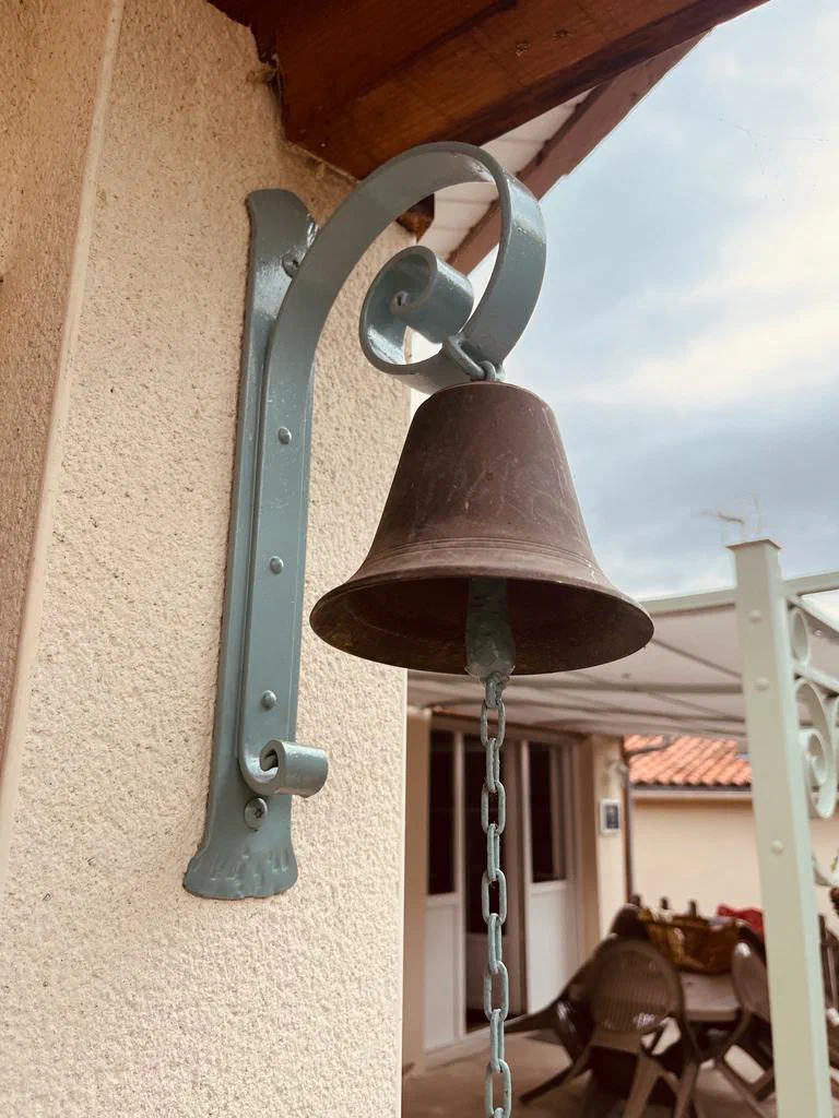 La cloche de bronze suspendue à sa potence de fer forgé, contre le mur de la maison