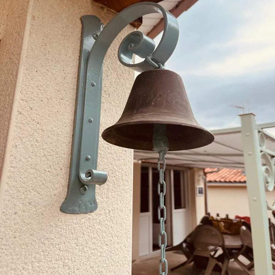 La cloche de bronze suspendue à sa potence de fer forgé, contre le mur de la maison