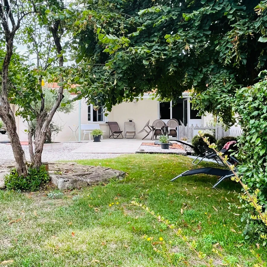La maison et sa terrasse vues du jardin, à travers les arbres, avec des transats sur la pelouse
