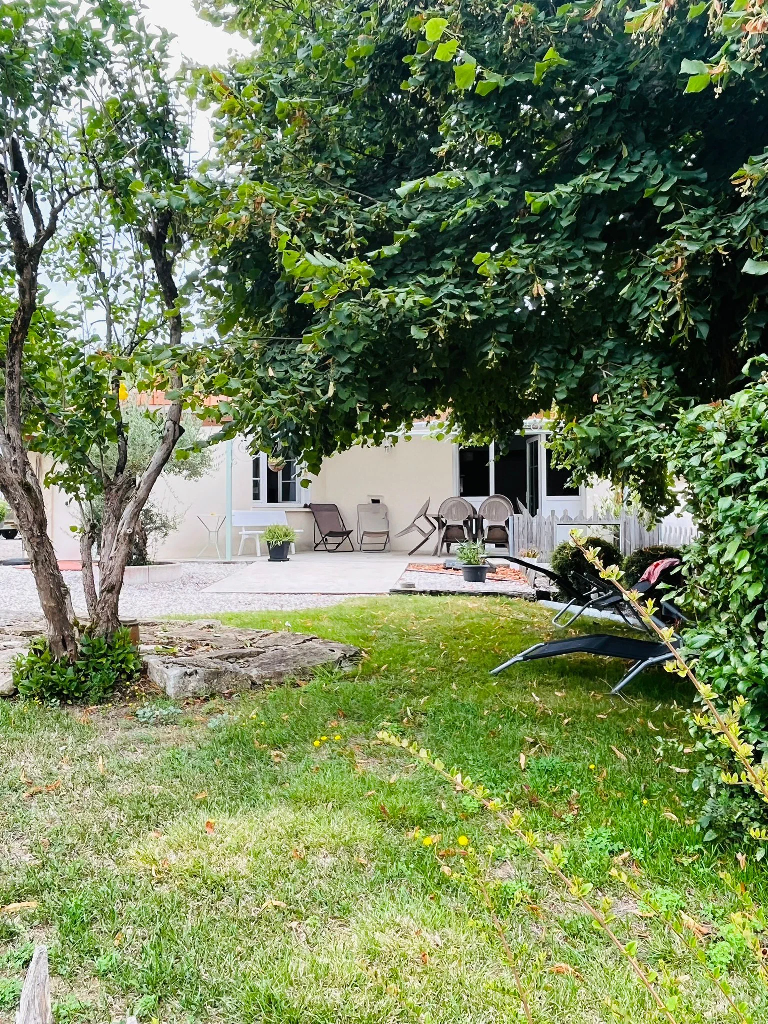 La maison et sa terrasse vues du jardin, à travers les arbres, avec des transats sur la pelouse