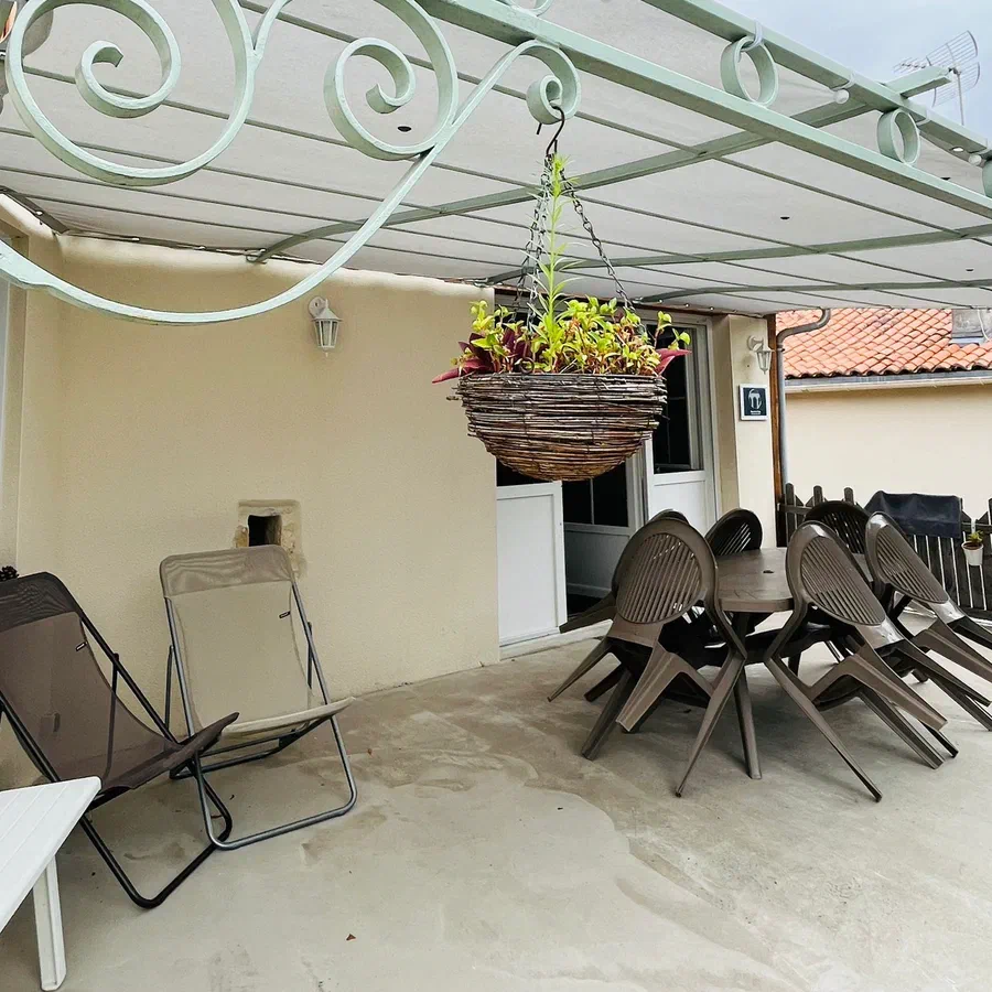 La terrasse abritée par une pergola en fer forgé, avec sa table, ses chaises et un panier suspendu
