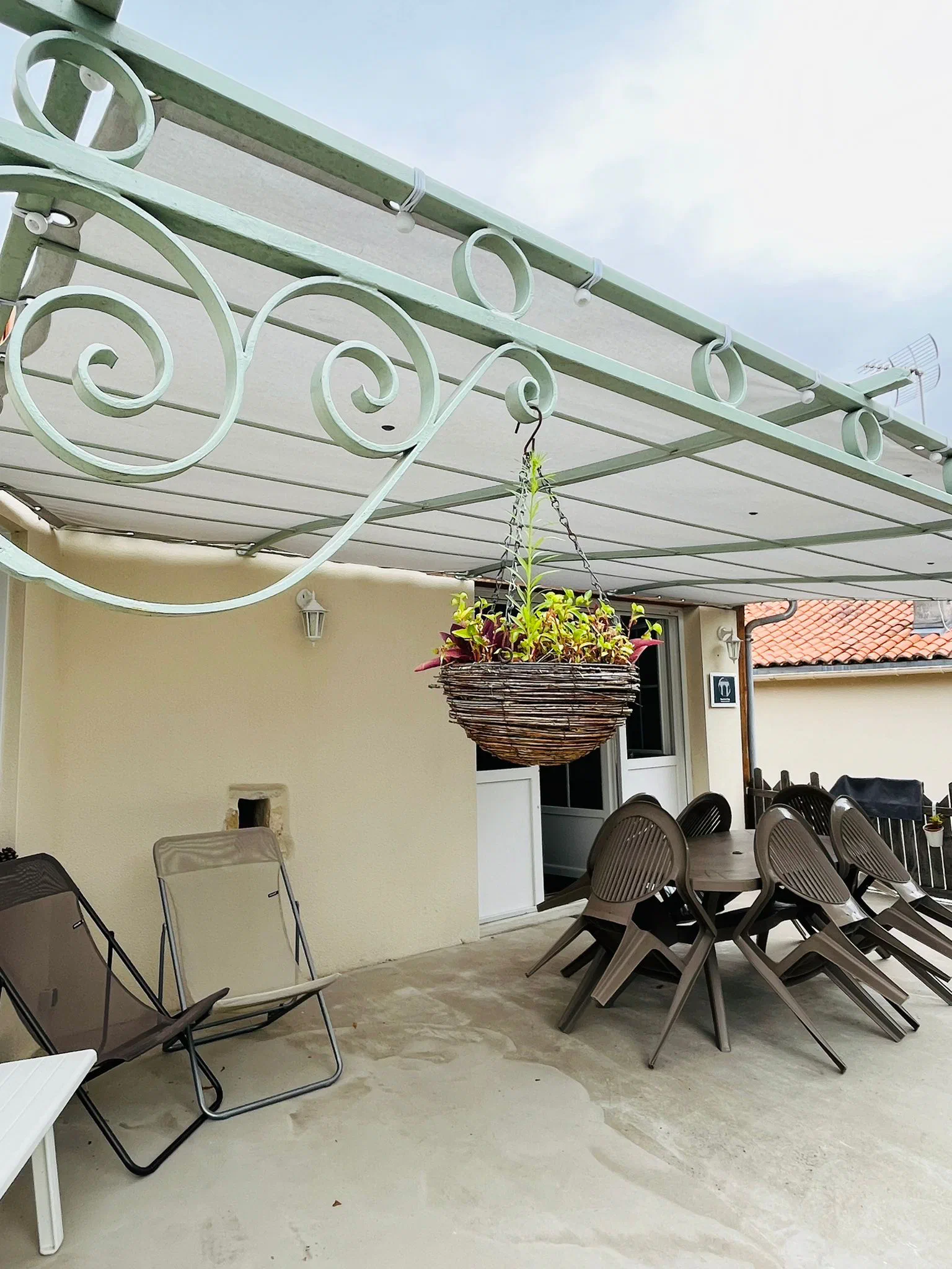 La terrasse abritée par une pergola en fer forgé, avec sa table, ses chaises et un panier suspendu